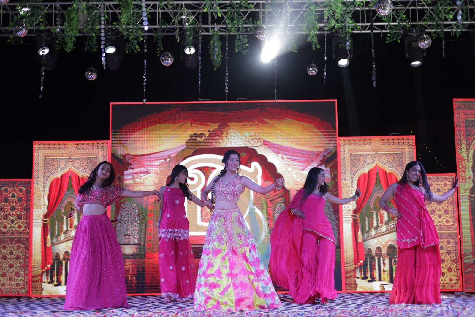 A group in pink lehengas holding the final pose on a lit wedding stage