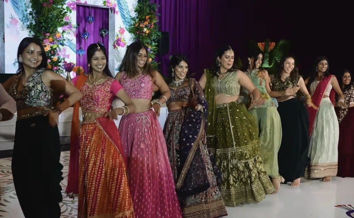 A line of women in lehengas dancing in front of a peacock-painted mandap