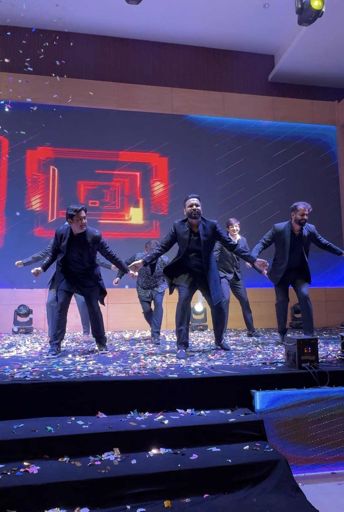 Five men in black holding a line across a lit stage under falling confetti
